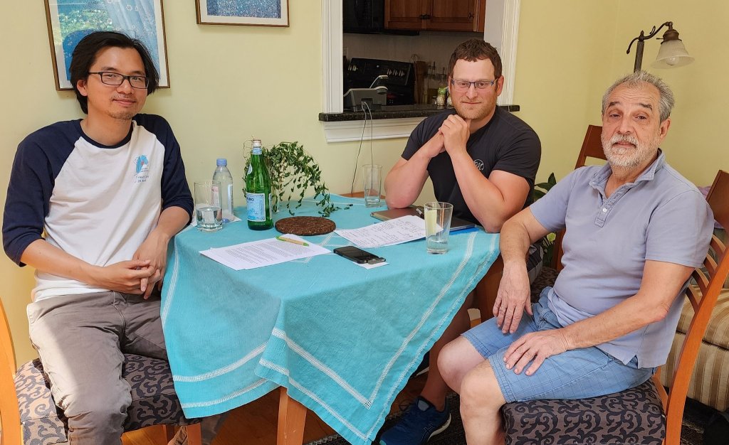 AMB CEO/CTO and Filamentech President working at a table with an off-center blue tablecloth and paperwork sitting in front of them, discussing their strategic partnership. They are wearing casual clothing and smiling at the camera. 