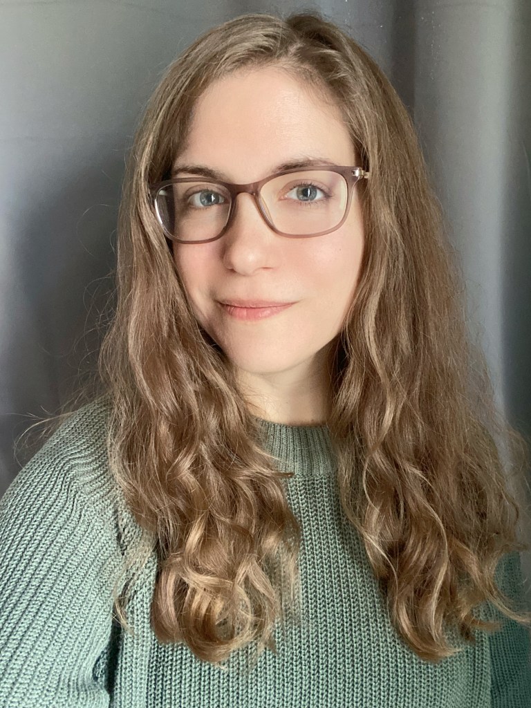 Portrait photo of a blonde woman in big glasses wearing a green knit sweater standing in front of a grey background.